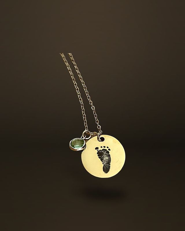 A gold-tone round disc pendant engraved with an infant footprint, hanging from a fine chain beside a small green stone charm, on a wooden surface with eucalyptus leaves.