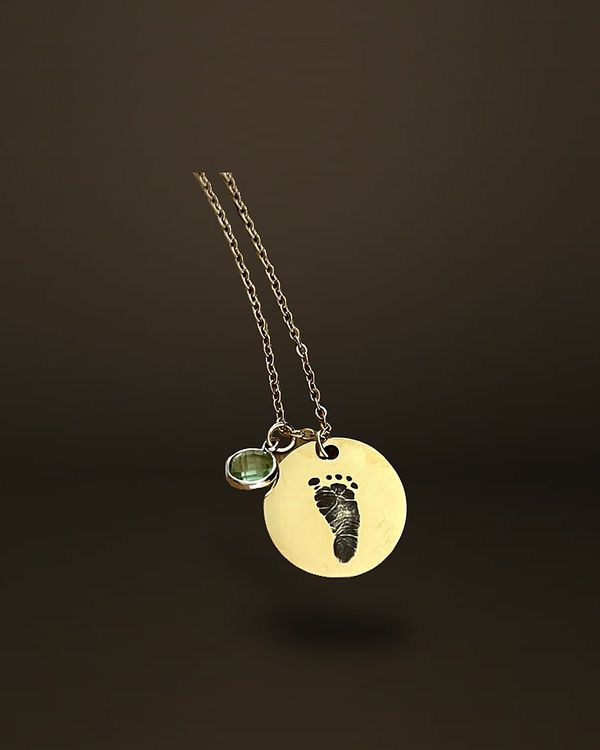 A gold-tone round disc pendant engraved with an infant footprint, hanging from a fine chain on a wooden surface.