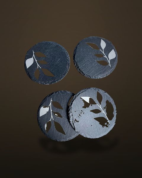 Four round slate coasters with irregular chipped edges, each engraved with a pale leafy-branch design, arranged on a warm off-white surface.