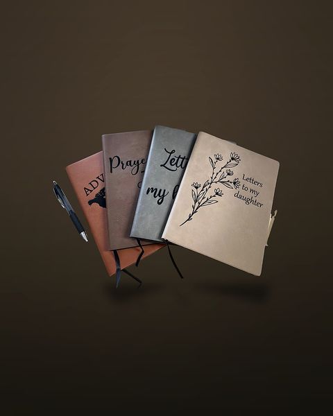 Four faux-leather journals fanned out on a beige countertop, engraved with different cover designs including a wildflower branch beside the words 'Letters to my daughter', with two pens alongside.