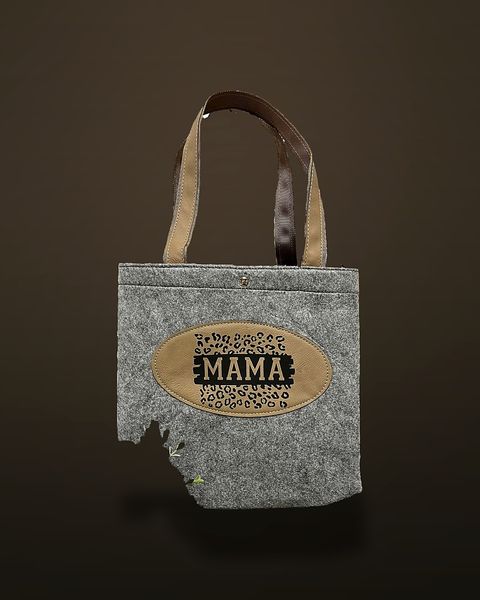 A grey felt tote bag with brown straps and a tan oval patch engraved 'MAMA' surrounded by a leopard-spot pattern, standing on a dark wood bench beside artificial greenery.